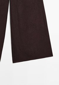 Pantalon marron foncé à jambes larges avec un détail en tissu texturé légèrement froissé sur fond blanc.