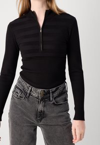 Schwarzes Oberteil mit Reißverschluss und horizontaler Streifenstruktur, kombiniert mit grauen, hoch taillierten Denim-Jeans mit Doppelknopfverschluss und leichtem Verblassen.