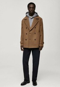Bruine double-breasted jas met omgeslagen revers, gedragen boven een grijze hoodie en zwarte broek, gecombineerd met zwarte schoenen.