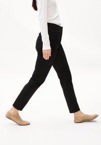 Pantalones de mezclilla negros con un corte ajustado, que cuentan con bolsillos traseros y una cintura de altura media, combinados con zapatos planos beige sobre un fondo liso.