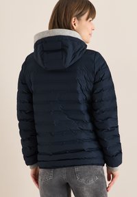 Veste matelassée bleu marine avec capuche et poignets gris, présentant un design matelassé horizontal. Le dos offre une coupe décontractée et une surface texturée.