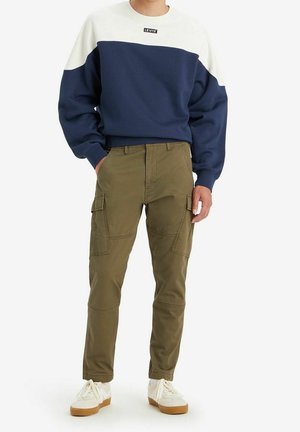 Mężczyzna w granatowo-białej bluzie Levi’s, oliwkowych spodniach typu cargo i białych trampkach z gumowymi podeszwami, stoi z rękami w kieszeniach.