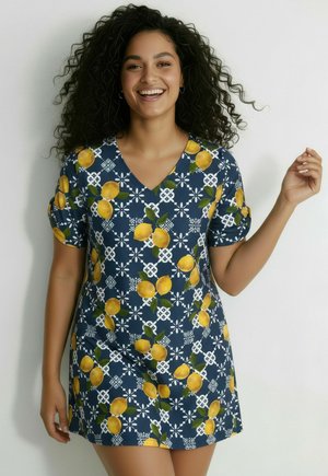 Femme souriante aux cheveux bouclés portant une robe bleu à encolure en V ornée de motifs de citrons jaunes et de formes géométriques blanches, posant devant un fond uni.