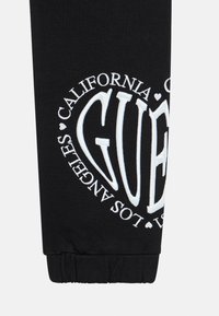 Svart joggebukse med elastisk mansjett, med stor hvit brodert tekst som leser "GUESS CALIFORNIA LOS ANGELES" i et sirkulært design.