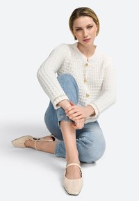 Cardigan tricoté blanc avec un col rond et quatre boutons dorés, associé à un jean bleu clair à revers et des ballerines beiges à bout pointu.
