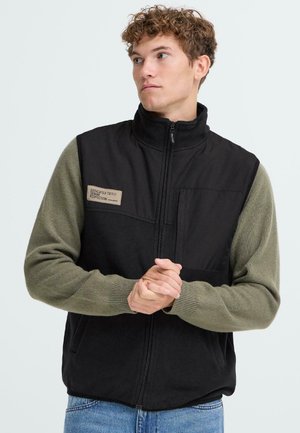 Gilet noir avec col montant et manches vertes, doté d'une poche avant et d'un patch de marque, fabriqué en tissu doux avec une finition mate.