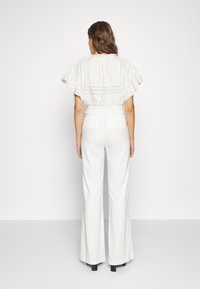 Weißbluse mit schwingenden Ärmeln und bestickten Details, kombiniert mit hochgeschnittenen weißen weit geschnittenen Hosen. Glatter Stoff, minimalistisches Design.