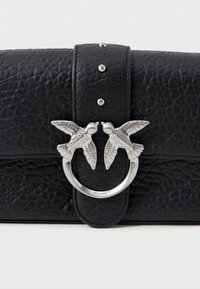 Melnas ādas somiņas ar teksturētu virsmu. Iekļauj sudraba apļveida aparatūru ar diviem detalizētiem putniem. Dekoratīvi piekariņi gar siksniņu.