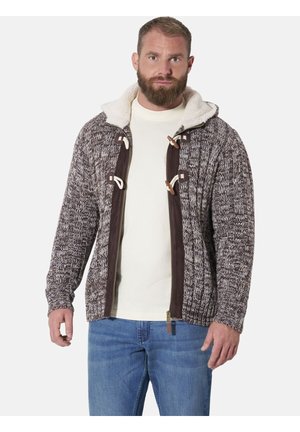 Man with beard wearing a brown and white knit hooded jacket with toggle buttons over a cream sweater and blue jeans, standing against white background.