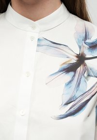 Κοντινή λήψη λευκού πουκαμίσου με μανταρινένιο γιακά και κουμπιά, που παρουσιάζει μεγάλο μπλε και μοβ λουλουδένιο σχέδιο στον δεξιό ώμο.