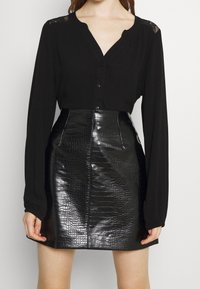 Blusa nera a maniche lunghe con un sottile dettaglio di pizzo sulle spalle, abbinata a una minigonna in faux pelle di coccodrillo nera con una cucitura frontale.