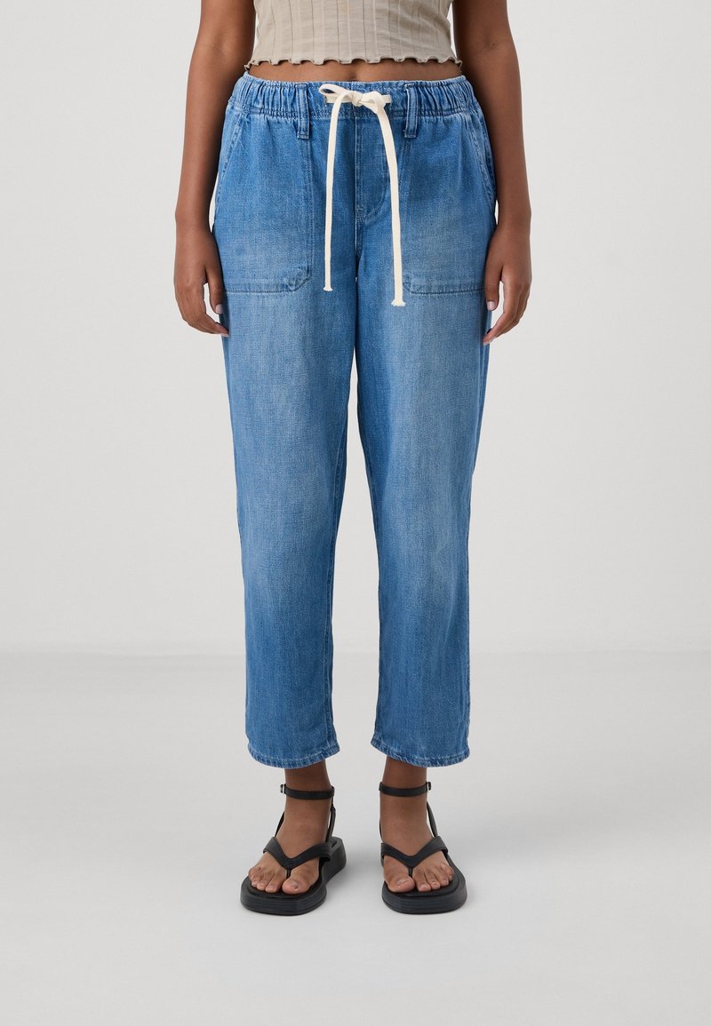 Jasnoniebieskie, luźne dżinsowe joggery z elastycznym pasem i białym sznurkiem. Zawierają dwie przednie kieszenie i skrócony dół.