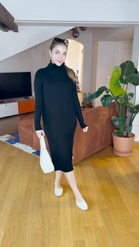 Μαύρο ριμενισμένο φόρεμα με μακριά μανίκια και ψηλό λαιμό, λευκές επίπεδες παπούτσια και λευκή τσάντα, στέκεται σε δωμάτιο με καφέ καναπέ και εσωτερικό φυτό.