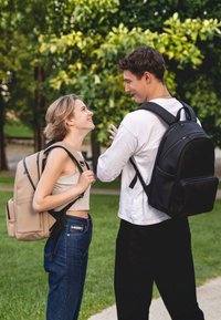 Beige és fekete hátizsákok cipzárral, párnázott pántokkal és szervező zsebekkel. Zöld lombozat előtt.