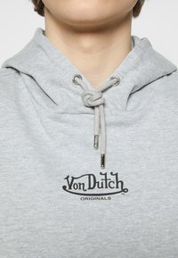 Personne portant un sweat à capuche gris clair avec le logo « Von Dutch Originals » imprimé sur la poitrine et des cordons noués.