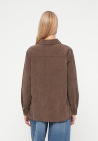 Blondyn stojący tyłem, ubrany w brązową koszulę z kordurowego materiału i niebieskie dżinsy na białym tle.