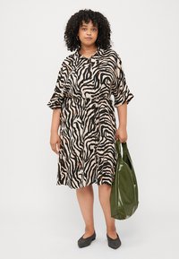 Švarko tipo suknelė smėlio spalvos su juodais zebra raštais, trumpomis rankovėmis, pabrėžta juosmeniu ir laisvu siluetu; derinama su dideliu žaliu blizgiu delniniu.