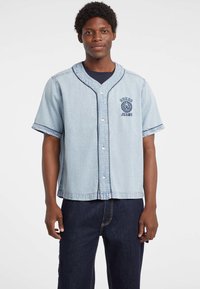 Lichtblauwe denim kortebloes met knoopsluiting aan de voorkant, contrasterende marineblauwe stiksels en geborduurd logo detail op de borst.