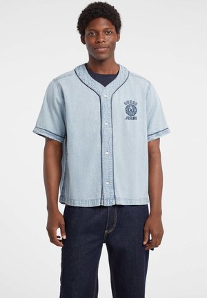 Lichtblauwe denim kortebloes met knoopsluiting aan de voorkant, contrasterende marineblauwe stiksels en geborduurd logo detail op de borst.