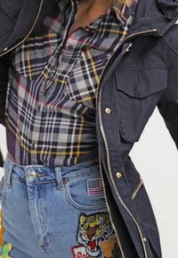 Navy parka with zippers, plaid shirt in gray, blue, and yellow, and high-waisted distressed denim shorts with embroidered patches.