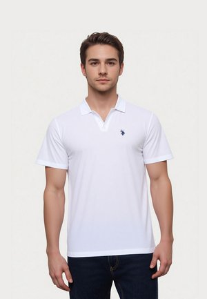 Tânăr care poartă un tricou polo alb simplu cu mâneci scurte și un mic logo închis la culoare pe pieptul stâng, stând în fața unui fundal deschis la culoare.