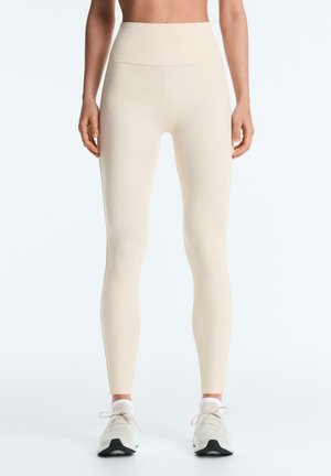 Žena nosiaca béžové legíny s vysokým pásom a biele bežecké topánky, stojaca s uvoľnenými rukami pri tele na bielom pozadí.