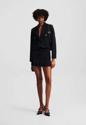 Blazer corto nero con due tasche anteriori, abbinato a una minigonna nera a balze e scarpe con tacco nere. Tessuto liscio e design su misura.