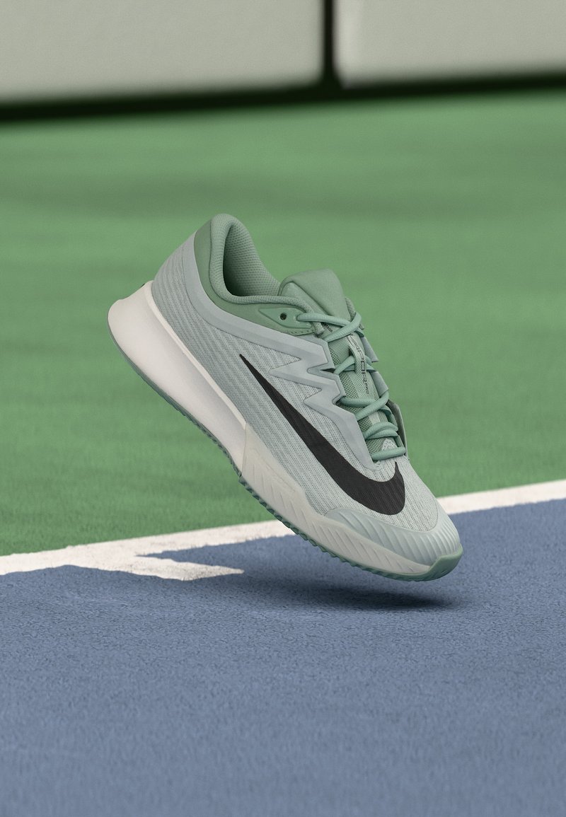 Ανοιχτό πράσινο παπούτσι τρεξίματος Nike με μαύρο λογότυπο swoosh, που αιωρείται πάνω από την επιφάνεια γηπέδου τένις σε μπλε και πράσινο κοντά στη γραμμή ορίου.