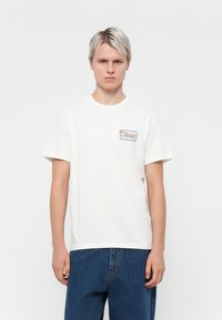 White cotton t-shirt with short sleeves, round neck, featuring a small brown and blue graphic logo on the left chest pocket area.