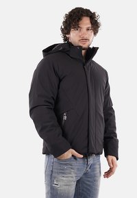 Uomo che indossa una giacca invernale nera con cappuccio e tasche con zip, insieme a jeans blu strappati, in piedi contro uno sfondo semplice.