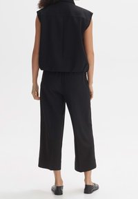 Czarny bezrękawnik z kołnierzykiem, zestawiony z czarnymi spodniami w wersji cropped. Materiał wydaje się gładki; projekt cechuje luźny krój. Czarne płaskie buty.