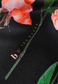 Čierna látka s jasným oranžovým kvetinovým potlačou, so zelenými listami a viditeľnou etiketou "by Ted Baker" v jemnom ružovom písmene.