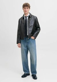 Braune Lederjacke mit spitzem Kragen, kombiniert mit hellblauen weit geschnittenen Jeans und schwarzen Schuhen. Schlichter Hintergrund.