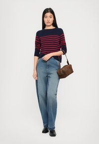 Maglione blu navy con strisce rosse, abbinato a jeans a gamba larga in blu chiaro a vita alta e una borsa in suede marrone. Scarpe nere completano il look.