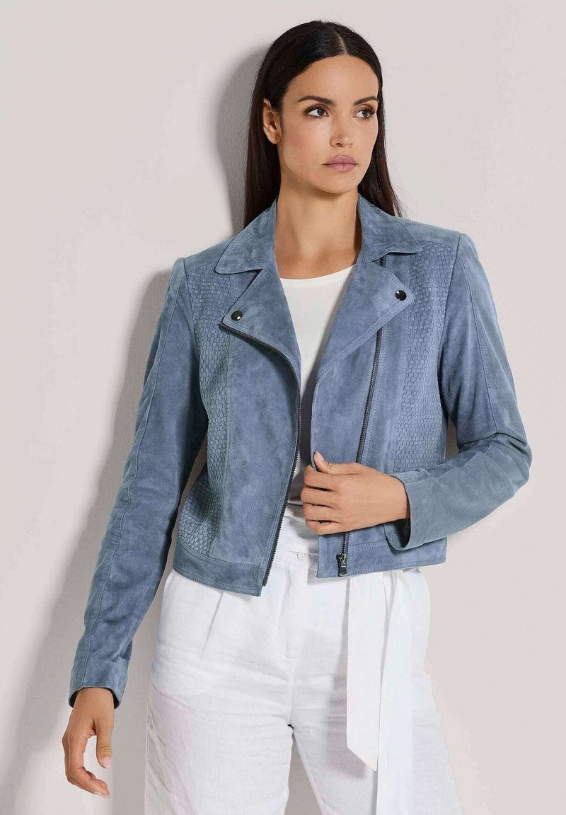 Frau in einer hellblauen Wildleder-Bikerjacke mit strukturierten Einsätzen und weißen hochtaillierten Hosen, die mit einem neutralen Hintergrund zur Seite blickt.