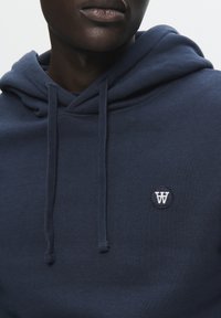 Mørkeblå sweatshirt med snor, blød overflade og et rundt broderet logo på brystet. Materialet ser ud til at være en blanding af bomuld.