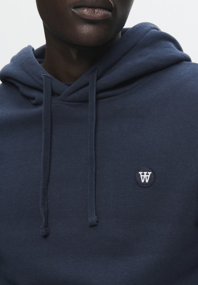 Haut à capuche bleu marine avec cordon de serrage, texture douce et logo brodé rond sur la poitrine. Le matériau semble être un mélange de coton.