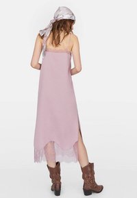 Langes, pastellpinkes Kleid mit verstellbaren Trägern und Spitzensaum, mit seitlichen Schlitzern. Kombiniert mit braunen Cowboy-Stiefeln und einem gemusterten Kopftuch.