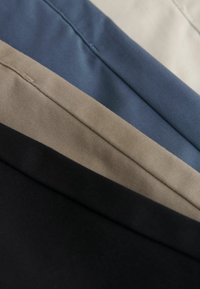 Quatre pièces de tissu pliées en noir, beige, bleu marine et écru, disposées en diagonale avec des coutures visibles et des détails de texture.