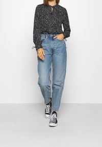 Blusa nera con motivo, maniche lunghe e polsini arricciati, abbinata a jeans a vita alta di colore blu chiaro e sneakers in tela nere.