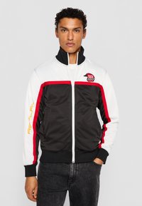 Black and white zip-up jacket with red accents, featuring a high collar, black ribbed cuffs, and a logo patch on the chest.