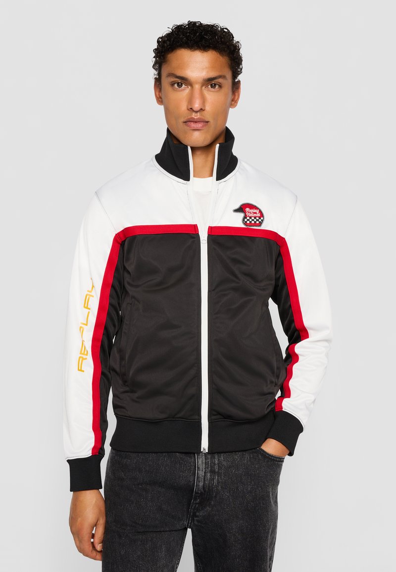 Schwarze und weiße Zip-Jacke mit roten Akzenten, ausgestattet mit einem hohen Kragen, schwarzen gerippten Bündchen und einem Logo-Patch auf der Brust.