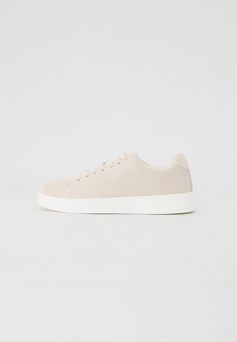 Beige semskinn sneakers med hvite gummisåler, avrundet tå og seks hull for snøring. Jevn tekstur med minimalistisk design.