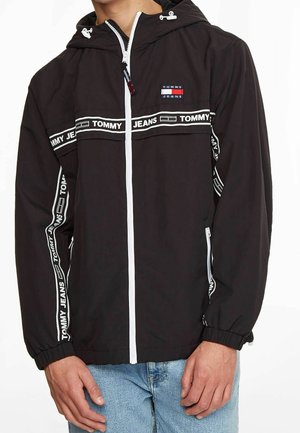 Veste à capuche noire zippée avec marquage blanc "Tommy Jeans" sur la poitrine, les manches et la fermeture éclair, portée sur un jean bleu clair.