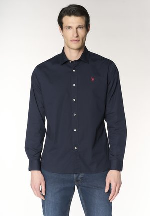 Uomo che indossa una camicia blu navy con bottoni e un logo rosso ricamato sul petto e jeans blu, in piedi davanti a uno sfondo semplice.