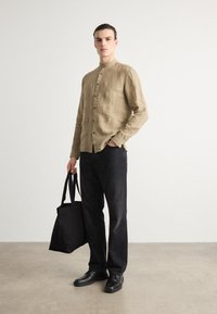Camisa de lino marrón claro con cuello mao y botones frontales, combinada con jeans oscuros y zapatos negros; sosteniendo una bolsa de lona negra.