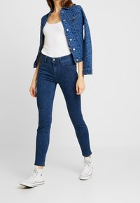 Denim jacket with a textured geometric pattern in dark blue, paired with matching fitted jeans and white sneakers.