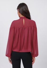 Bordeauxfarbige Bluse mit langen, puffigen Ärmeln, einem hohen Kragen und einem Schlüssellochdetail auf der Rückseite. Hergestellt aus leichtem, durchsichtigem Stoff.