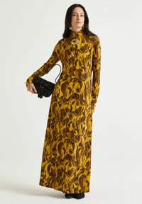 Maxikleid aus gelbem Stoff mit floralem braunem Muster, hohem Kragen, langen Ärmeln und figurbetonter Silhouette. Kombiniert mit einer schwarzen Handtasche.