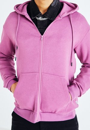 Person trägt einen rosa Kapuzenpullover mit Reißverschluss und Fronttaschen über einem schwarzen Shirt, Hände in den Kapuzenpockets, sichtbare Halskette mit Kette.